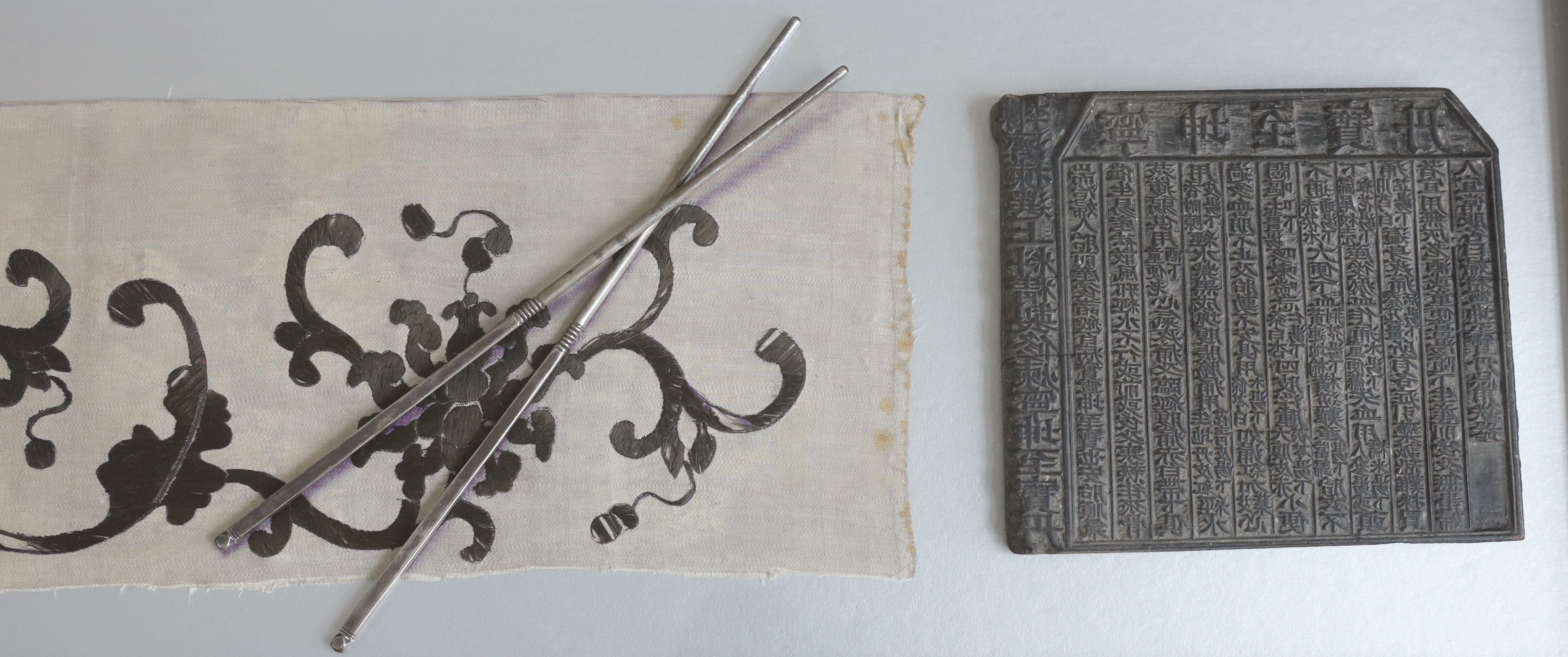 A late 19th century Chinese black and cream silk embroidery, mounted together with an inscribed calligraphic stone, a pair of white metal chop sticks and a similar dish, embroidery: 5.5cm wide, 105cm long